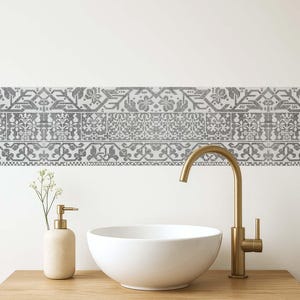 Può includere: Un mobile da bagno con un lavabo in ceramica bianca, un rubinetto dorato e un dosatore di sapone beige. Il mobile in legno ha un bordo decorativo di piastrelle a motivi grigi e bianchi. Un piccolo rametto di fiori bianchi.