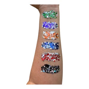 May include: A close-up of a person's arm with six different swatches of glitter. The swatches are in the following order: green, purple, orange, blue, red, and black.