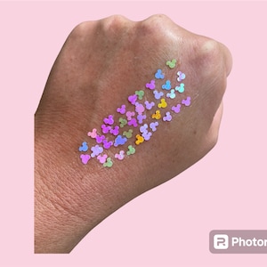 May include: A hand with a scattering of iridescent, multi-colored Mickey Mouse head shaped sequins. The sequins are in shades of pink, purple, green, yellow, and blue.
