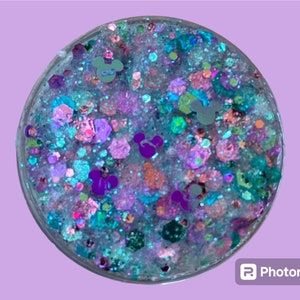 May include: A round, clear container filled with colorful glitter and small Mickey Mouse-shaped confetti. The glitter includes shades of blue, purple, pink, and green, creating a sparkling effect. The container is set against a light purple background.
