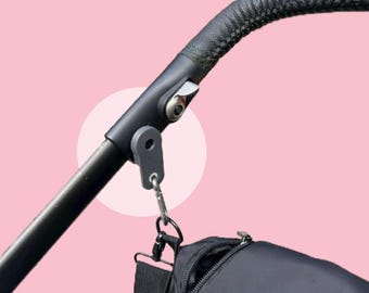 May include: A black pushchair handle with a textured grip and a metal clasp. A black bag is attached to the handle with a metal carabiner. The background is a soft pink.