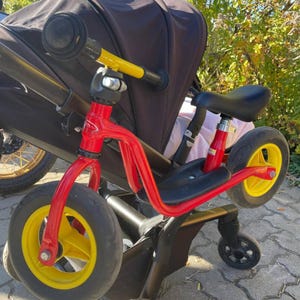 Könnte beinhalten: Ein rotes und gelbes Laufrad mit schwarzen Reifen und einem schwarzen Sitz. Das Fahrrad hat einen gelben Lenker und steht neben einem schwarzen Kinderwagen. Das Fahrrad ist für kleine Kinder konzipiert.