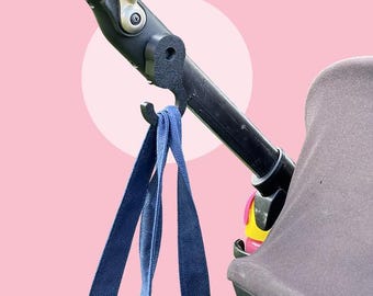 May include: A black stroller hook supporting a navy blue tote bag. The hook is attached to the stroller handle, with a portion of the stroller's canopy visible. The background is a light pink with a white circle.