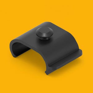 Può includere: Una clip metallica nera e curva con una parte superiore arrotondata e una piccola testa a vite circolare. La clip ha una finitura liscia e opaca ed è posta su uno sfondo giallo. Il design della clip suggerisce che viene utilizzata per fissare o tenere oggetti.