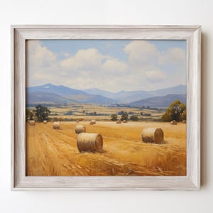 Op de afbeelding: Een ingelijst landschapsschilderij dat een landelijke scène weergeeft met gouden hooibalen in een veld. Het kunstwerk toont een lichtblauwe lucht met pluizige witte wolken, glooiende heuvels en verre bergen. De lijst is licht en neutraal.