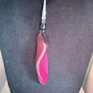 May include: A pink and black feather charm with a black bead and silver clip. The charm is hanging from a black cord.