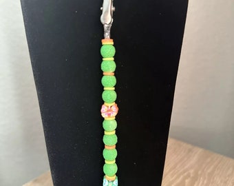 May include: A green beaded lanyard with a silver clip and colorful butterfly charms. The lanyard is made of green, orange, and yellow beads with a pink and white flower charm.