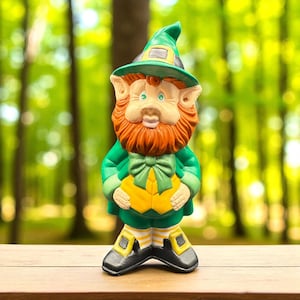 May include: A ceramic leprechaun figurine, featuring a green hat and coat, a red beard, and a yellow vest. The figurine has black shoes with yellow buckles and is standing on a wooden surface. The background is a blurred view of a green forest.