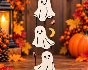 Può includere: Una decorazione verticale da appendere composta da tre sagome di fantasmi in legno bianco con tratti del viso e dettagli neri, collegati da uno spago nero e arricchiti da una piccola luna crescente gialla e pipistrelli marroni.
