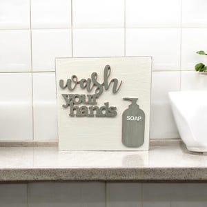 May include: White wooden sign with the words "wash your hands" in green letters and a green silhouette of a soap dispenser with the word "soap" on it.