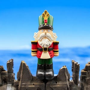 May include: A red, green, and white nutcracker figurine stands on a weathered, gray surface. The nutcracker has a black hat with a gold plume, a white beard, and a black mustache. The background is a blue sky with white clouds.