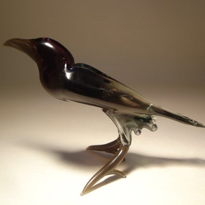 May include: A glass crow figurine with a dark purple head and black body. The crow has a long, thin tail and stands on a brown glass base.