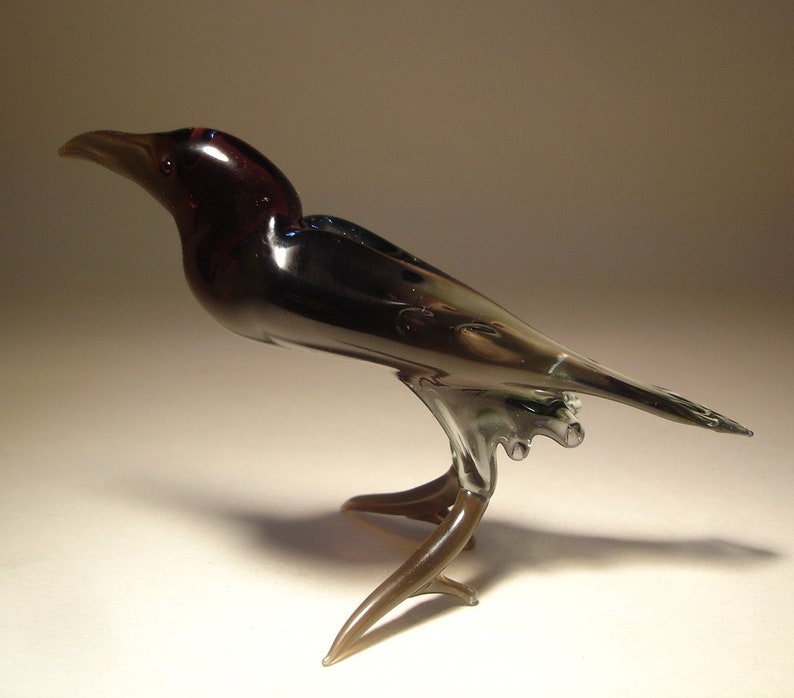 May include: A glass crow figurine with a dark purple head and black body. The crow has a long, thin tail and stands on a brown glass base.
