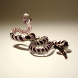 Peut inclure: Une figurine de serpent en verre avec un corps enroulé et une tête relevée. Le serpent est transparent avec des bandes alternées de marron foncé et de violet clair. La tête est violet clair et blanche.