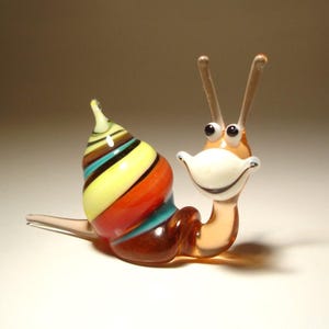 May include: A glass snail figurine featuring a multi-colored spiral shell with horizontal bands of yellow, orange, teal, and dark brown. The body is a translucent light brown with two long antennae, black eyes, and a wide, white, smiling mouth.