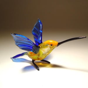 Pode incluir: Uma estatueta de beija-flor em vidro com asas azuis translúcidas, cabeça amarelo-brilhante e corpo multicolorido com manchas amarelas e laranjas. O pássaro possui um bico longo, fino e escuro e pernas pretas delgadas, em pose de voo.
