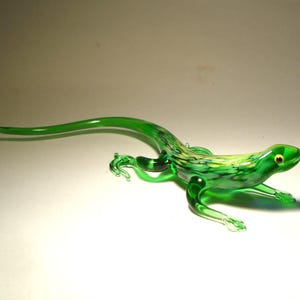 Puede incluir: Figura de lagarto de vidrio hecha a mano en tonos verdes, con detalles amarillos y negros. El lagarto tiene una cola larga y curvada y patas detalladas, y mide aproximadamente 20 cm de largo.