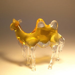 May include: A glass camel figurine with a clear body and amber coloured hump. The figurine is standing on a light brown surface.