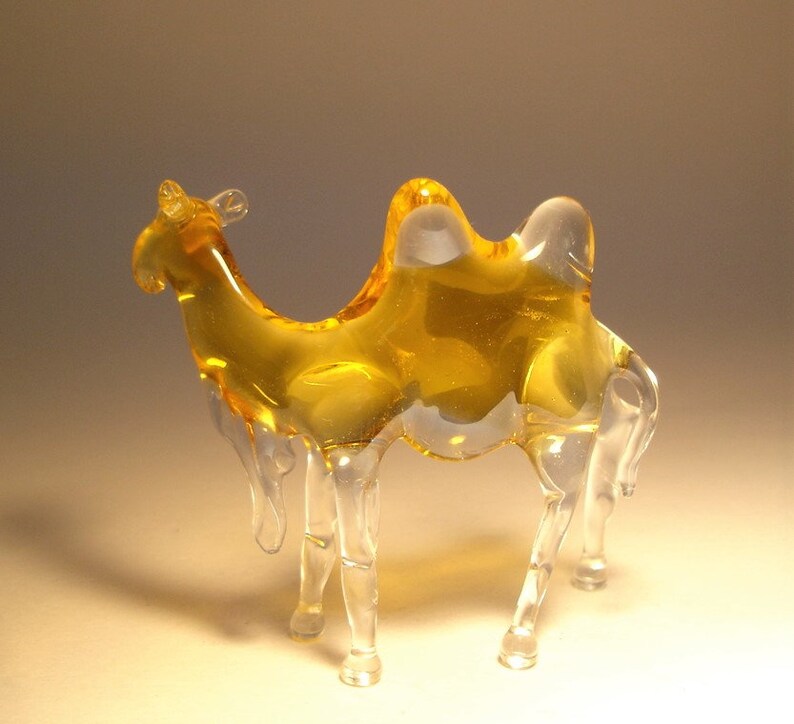 May include: A glass camel figurine with a clear body and amber coloured hump. The figurine is standing on a light brown surface.