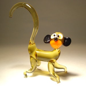Könnte beinhalten: Eine Glasfigur eines Affen mit einem langen, gekräuselten Schwanz. Der Affe ist gelb mit schwarzen Ohren und einem leuchtend orangefarbenen Mund.