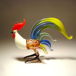 May include: A glass rooster figurine featuring a white body, a bright red comb and wattle, and a multi-colored tail with long, flowing yellow, blue, and green glass plumes. The rooster has a speckled brown midsection and thin black glass legs.
