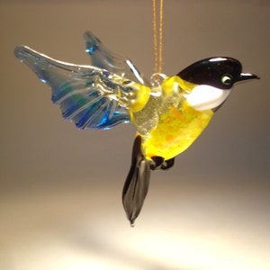 May include: A glass bird ornament with outstretched wings. The bird features a black head, white chest, and yellow body with orange speckles. Its wings are transparent blue and green. A gold string is attached for hanging.