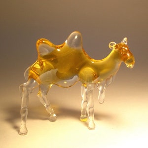 May include: A glass camel figurine with a clear body and a yellow hump. The figurine is standing on a white surface.