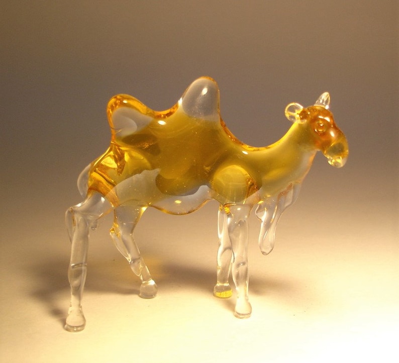 May include: A glass camel figurine with a clear body and a yellow hump. The figurine is standing on a white surface.