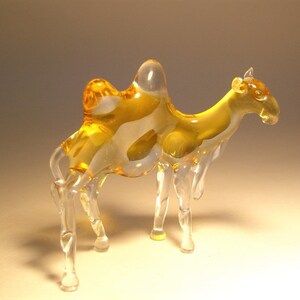 May include: A glass camel figurine with a clear body and amber coloured humps. The figurine has a detailed design and is a decorative piece.