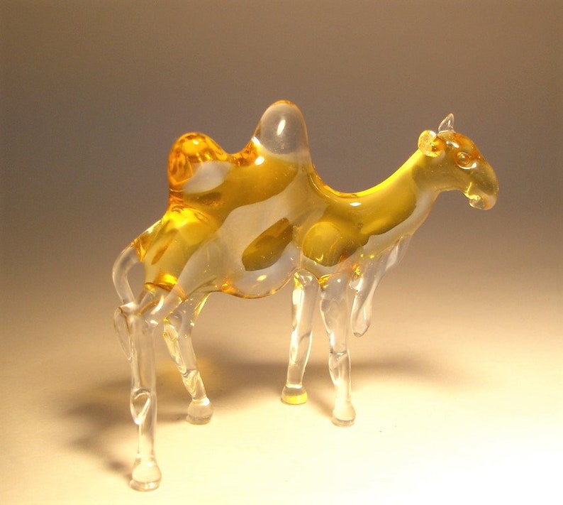 May include: A glass camel figurine with a clear body and amber coloured humps. The figurine has a detailed design and is a decorative piece.