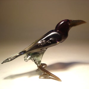 May include: A glass crow figurine with iridescent purple and black feathers. The crow is perched on a branch with its wings spread out.