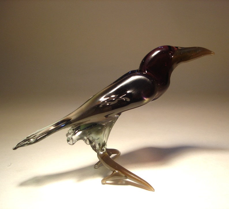 May include: A glass crow figurine with iridescent purple and black feathers. The crow is perched on a branch with its wings spread out.