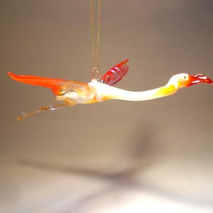 May include: A glass ornament shaped like a flamingo with orange and white wings and a yellow beak. The ornament is hanging from a gold string.