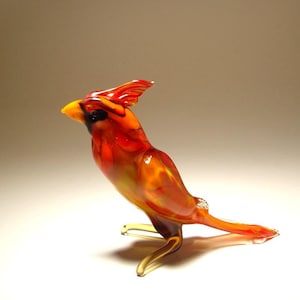 May include: A glass cardinal figurine in shades of red, orange, and yellow. The bird has a yellow beak and a crest. The figurine is posed in a standing position, with one leg extended. The glass is translucent.