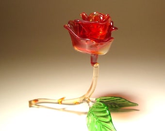 May include: A handcrafted glass rose sculpture. The rose is a vibrant red color, with a clear stem and a green leaf. The glass is translucent, allowing light to pass through. The sculpture is a decorative item.