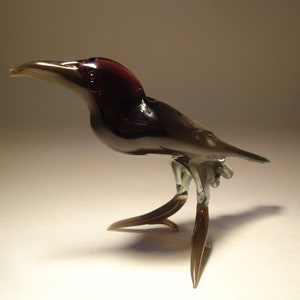 May include: A small glass bird figurine with a dark purple head and black body. The bird has long, thin legs and a long, thin beak. The bird is standing on a white surface.