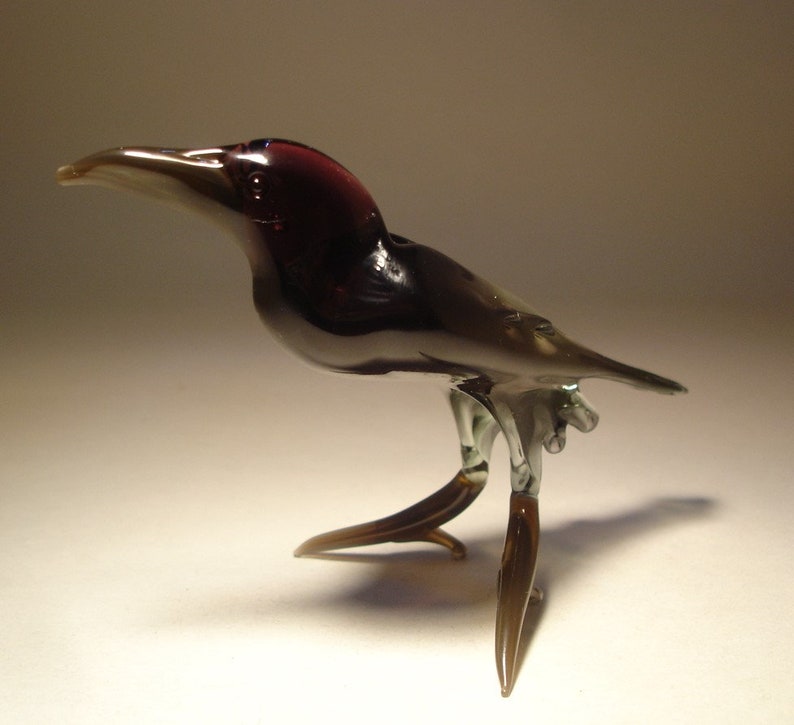 May include: A small glass bird figurine with a dark purple head and black body. The bird has long, thin legs and a long, thin beak. The bird is standing on a white surface.