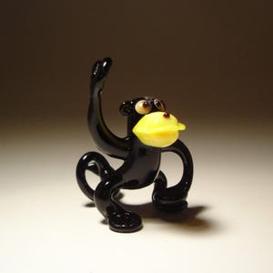 May include: A small, handcrafted glass monkey figurine. The monkey is black with a yellow face and large, round eyes. Its arm is raised, and it has a curled tail. The figurine is displayed on a white surface.