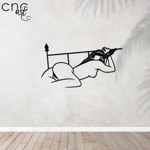 以下が含まれることがあります： ベッドに横たわる女性の白黒の線画の壁飾り。女性はリラックスした姿勢で、頭を手に乗せている。ベッドはヘッドボードとフットボードが付いたシンプルなデザインです。