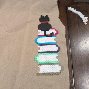 Pode incluir: Uma figura de gato colorida e pixelizada feita de contas de plástico fundidas. O gato preto com uma coleira vermelha está empoleirado em cima de uma pilha de livros de várias cores, incluindo azul, roxo e verde. O projeto de artesanato está em papel castanho.