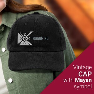 Puede incluir: Gorra de béisbol de pana negra con un símbolo maya bordado en blanco y el texto "Hunab Ku".