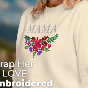 Puede incluir: Una sudadera de color crema con la palabra "MAMA" bordada en hilo plateado. Debajo de la palabra, se borda un diseño floral con flores rosas, moradas y verdes. El texto "Wrap Her In LOVE Embroidered Sweatshirt FOR MOM" está impreso debajo del diseño.