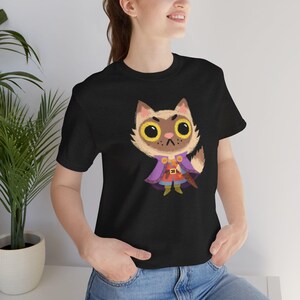 May include: Black t-shirt featuring a cartoon cat character wearing a purple cape and orange tunic. The cat has large yellow eyes and a stern expression. The design is centered on the shirt, creating a playful and whimsical aesthetic.
