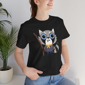 May include: Black t-shirt featuring a cartoon raccoon character with large blue eyes, a yellow scarf, and a wooden stick. The raccoon is wearing a blue outfit. The design is a whimsical illustration, perfect for casual wear.