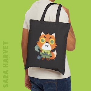 May include: Black tote bag featuring a cartoon fox illustration. The fox is orange and white with large yellow eyes, wearing a green scarf and holding a blue object. The text "SARA HARVEY" is printed vertically on the left side of the bag.