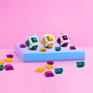 May include: Three geometric puzzle cubes with white, teal, yellow, and purple accents sit on a blue platform. Scattered around are teal, yellow, and purple geometric shapes. The background is a solid pink.