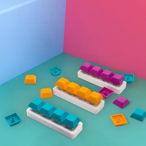 May include: Three sets of keyboard keycaps are shown, each with four keys in blue, yellow, and purple, mounted on white bases. Loose keycaps in matching colours are scattered on a teal surface. The background is a gradient of blue and pink.