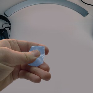 Può includere: Una mano che tiene una pietra blu chiaro di forma irregolare. La pietra è traslucida e sembra essere un cristallo o un minerale lucido. Lo sfondo include una superficie bianca, un paio di cuffie nere e un monitor del computer.