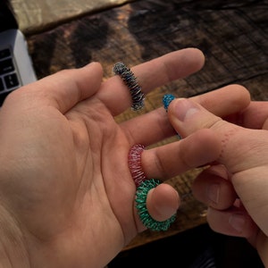 Pode incluir: Um close-up de mãos segurando e usando vários anéis de massagem para os dedos coloridos. Os anéis são feitos de metal enrolado em várias cores, incluindo prata, azul, rosa e verde. O fundo inclui uma superfície de madeira e um laptop.