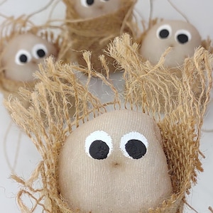 May include: Four handmade, beige-colored bird ornaments with large, painted black eyes and white highlights. Each bird is nestled in a nest made of woven, natural-colored twine, creating a rustic, whimsical aesthetic.